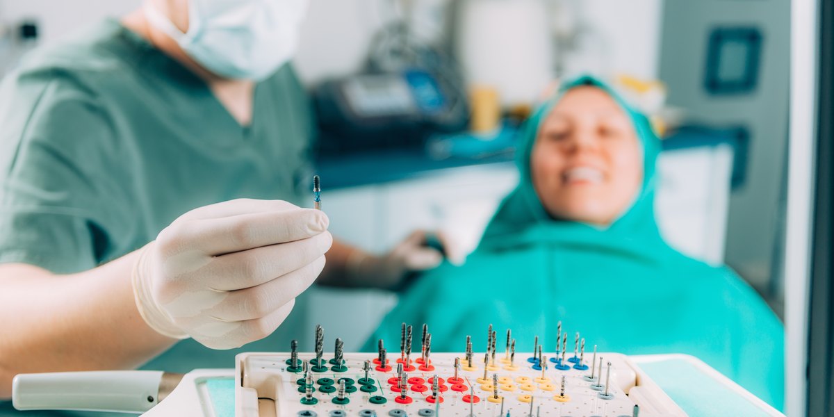 Implante dental: en el quirófano, la mano enguantada del dentista saca una fresa del kit de cirugía de implantes mientras la paciente espera tranquila en el sillón