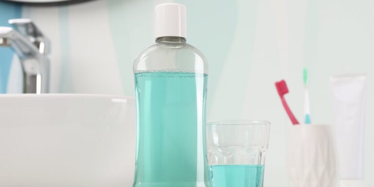 Enjuague bucal con clorhexidina: botella de enjuague bucal, cepillos de dientes y vidrio sobre mesa blanca en el baño