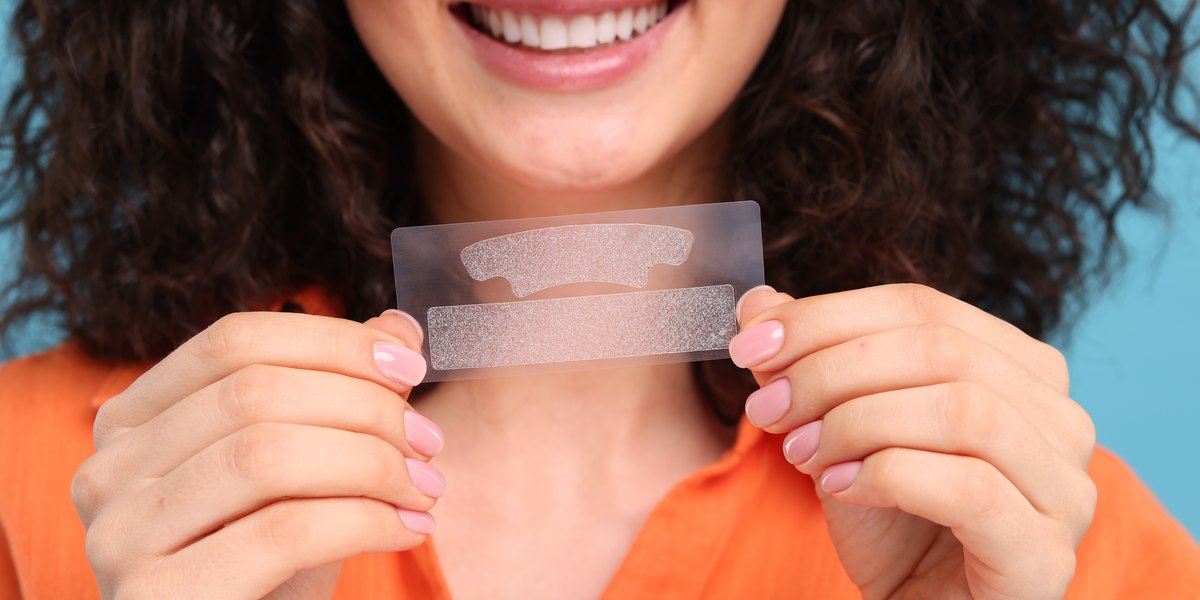 Mujer joven que sostiene tiras blanqueadoras de dientes sobre fondo azul claro, primer plano